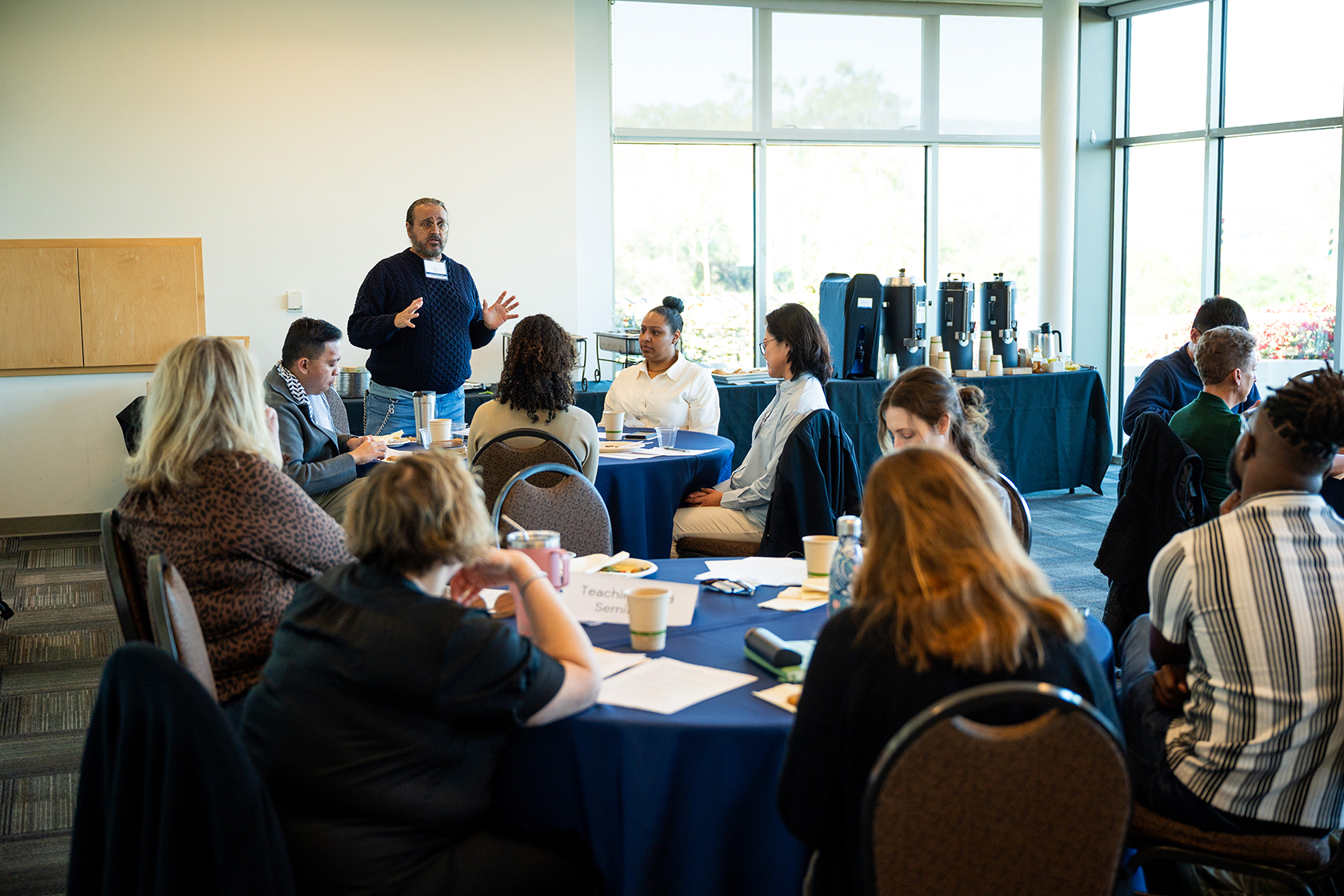 room of people seated around circular tables one person standing in the background
