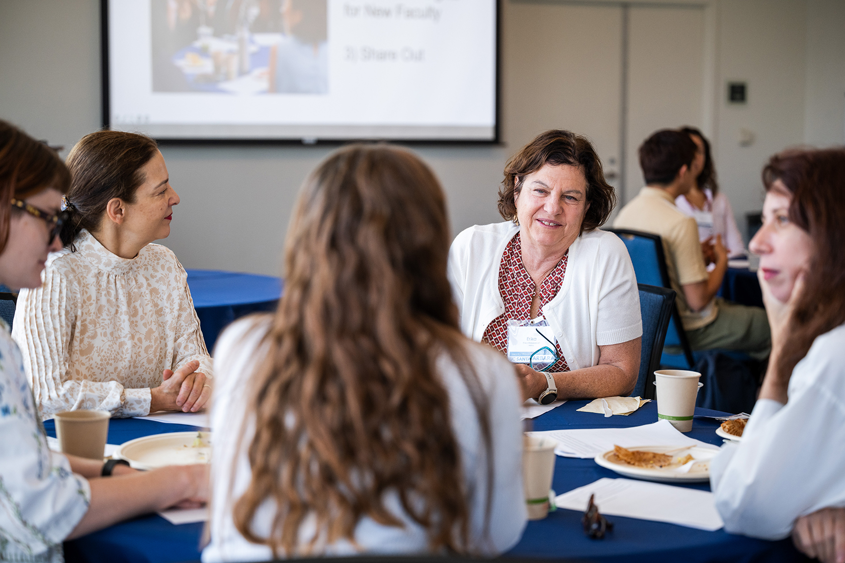 Attendees sit around blue covered table engaged in conversation with a projector in the background