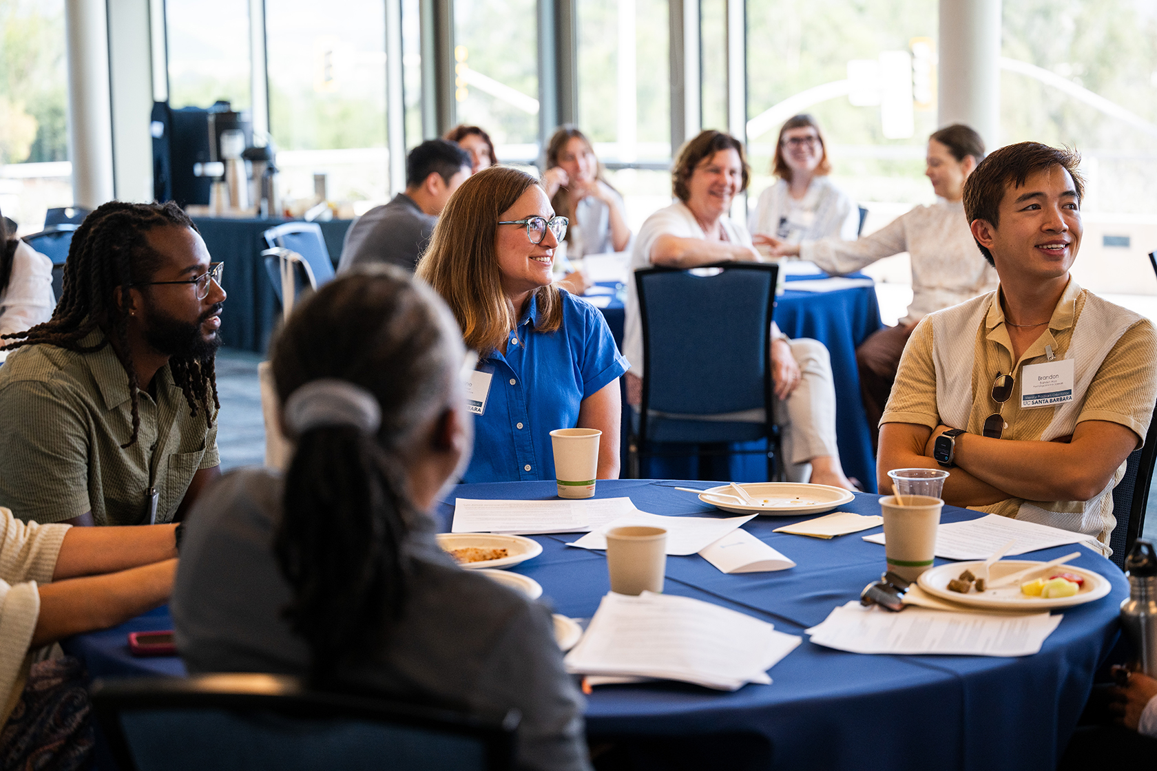 Group of attendees sit at round table smiling while listening to speaker