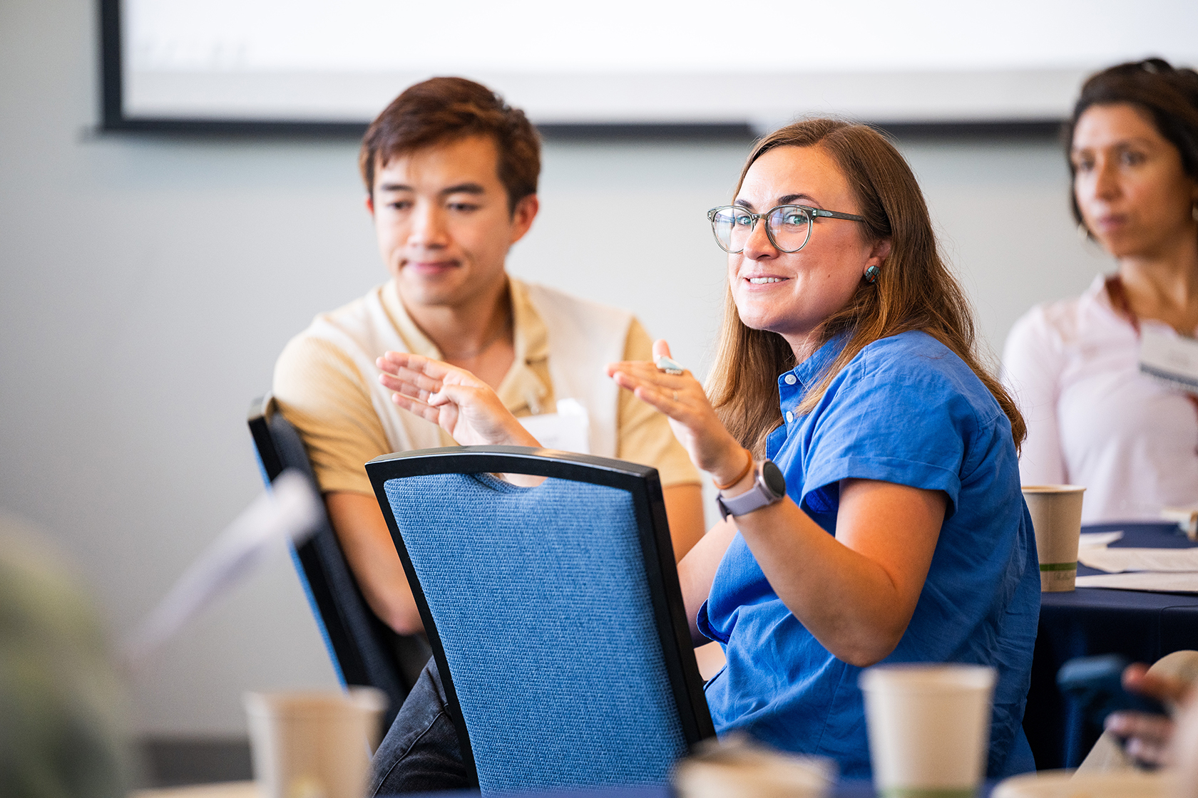 Speaker in blue blouse and glasses speaks and gestures during a discussion as other attendees are seated nearby