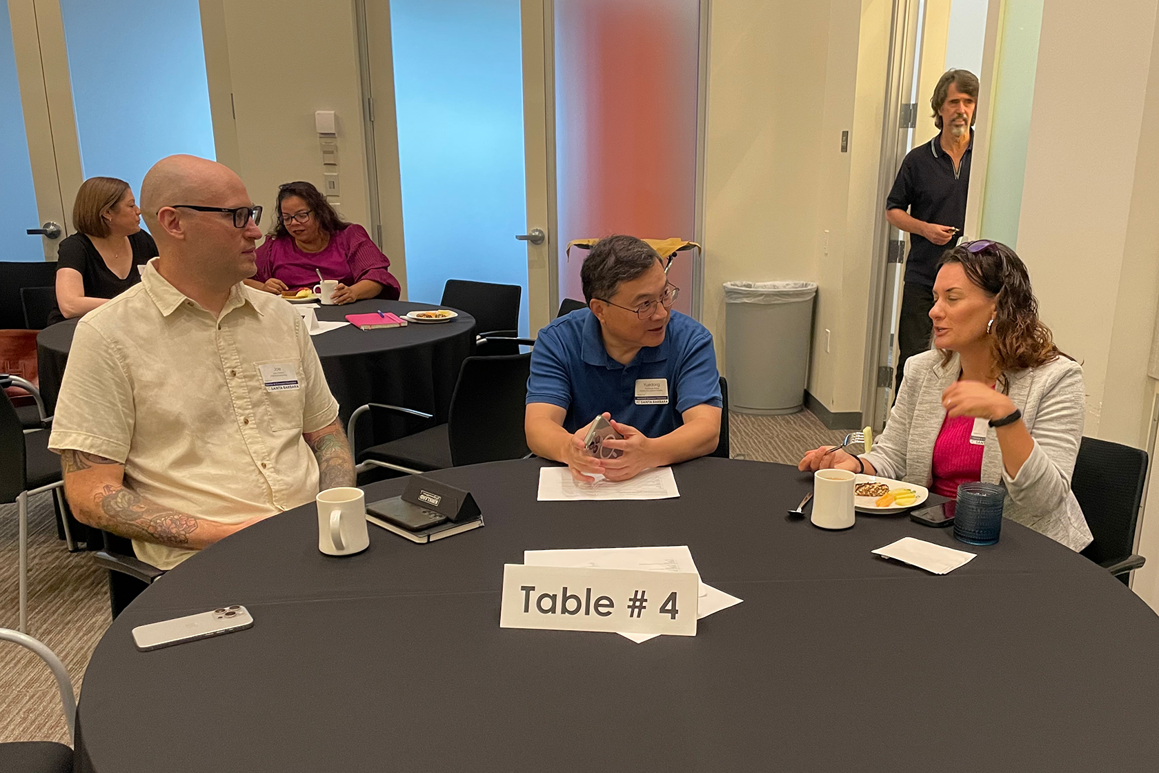 two circular tables with people seated and talking one person standing in a door frame in the background