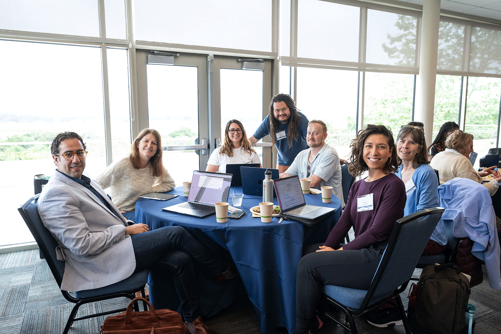 six people seated one person standing around a circular table with a blue tablecloth