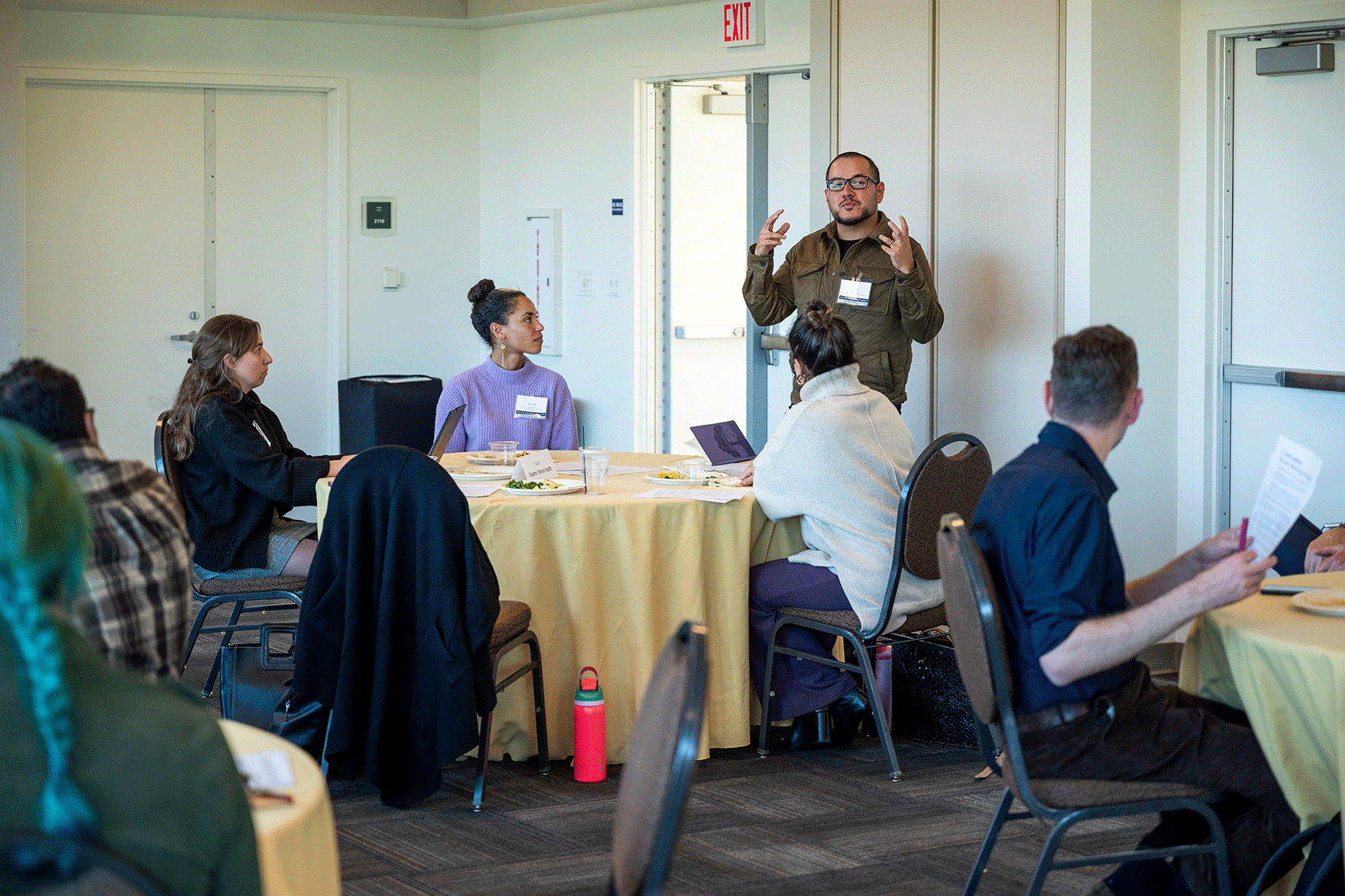six people seated around circular tables one person standing talking