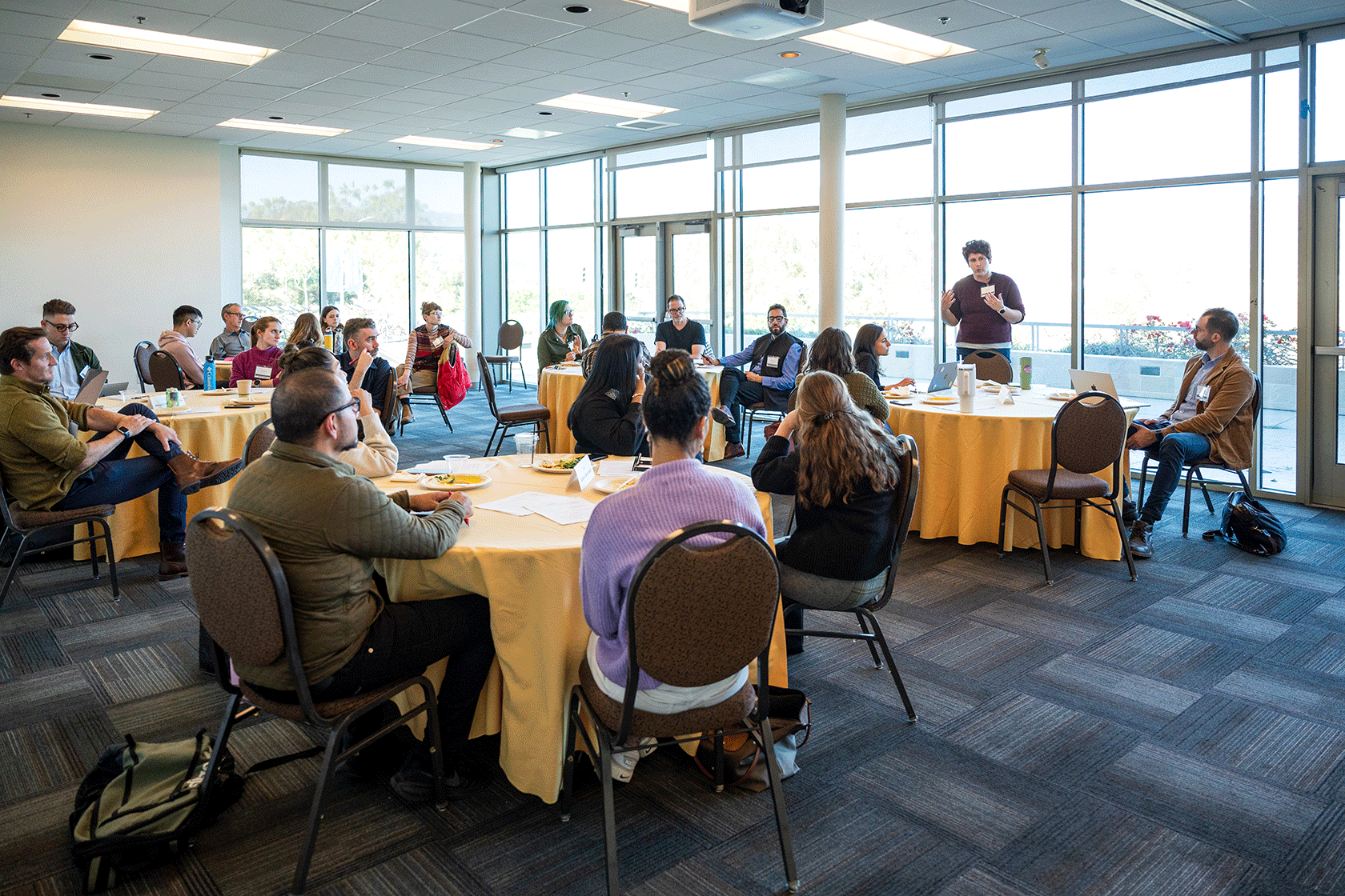 conference room with several circular tables people seated around them and one person standing talking