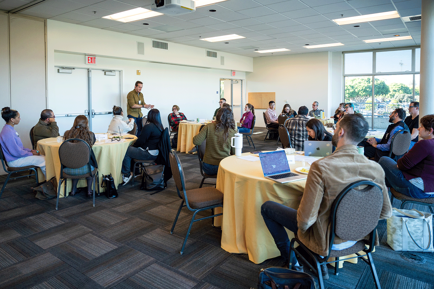 large group of people seated around circular tables with one person standing and speaking