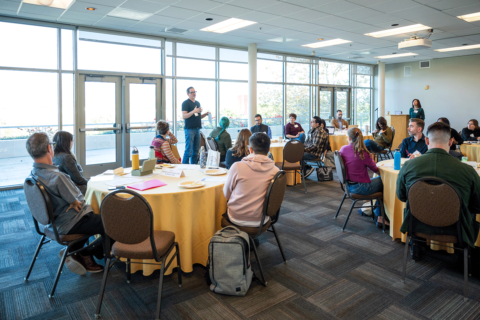 conference room with several circular tables people seated around them and one person standing talking one standing at a podium