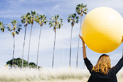 Image of UCSB Coast with woman holding yellow exercise ball, with palm trees in the background